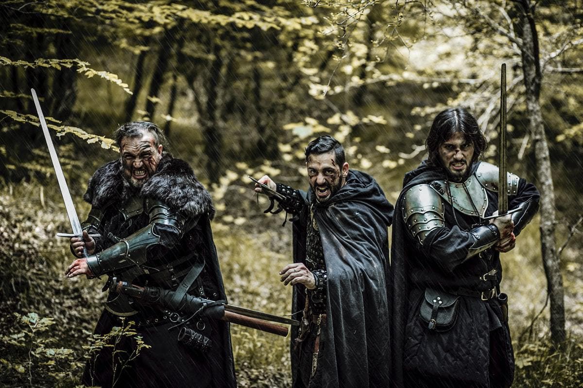 three men dressed in cloaks and armor, holding swords aggressively in the woods