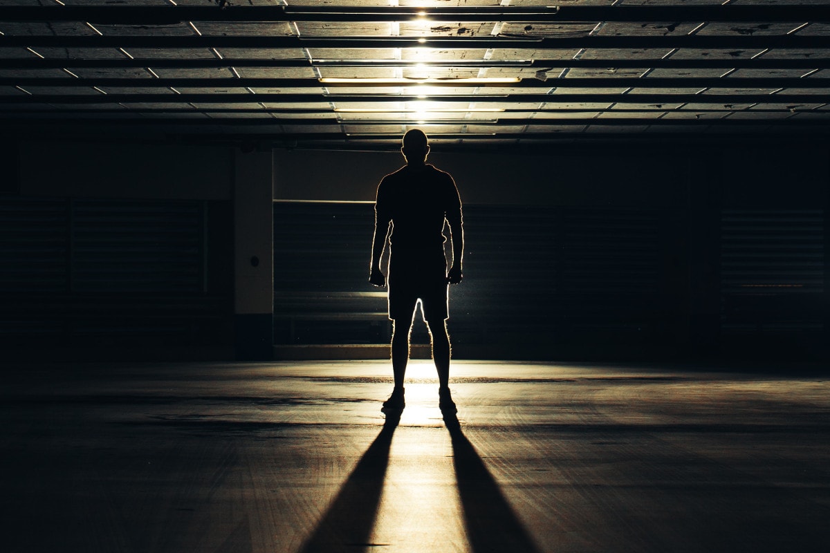 A man standing in a large, dark room with a light behind him. He is silhouetted.